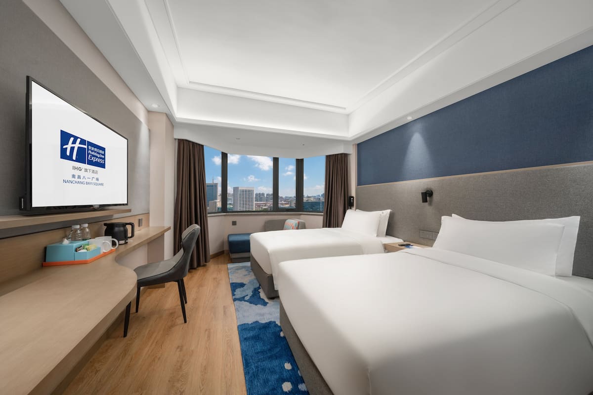 A hotel room with two beds, a desk, a chair, a TV, and a city view.