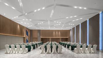 A conference room with rows of tables and chairs, a high ceiling with recessed lighting, and large windows on one side.