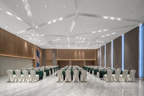 A conference room with rows of tables and chairs, a high ceiling with recessed lighting, and large windows on one side.