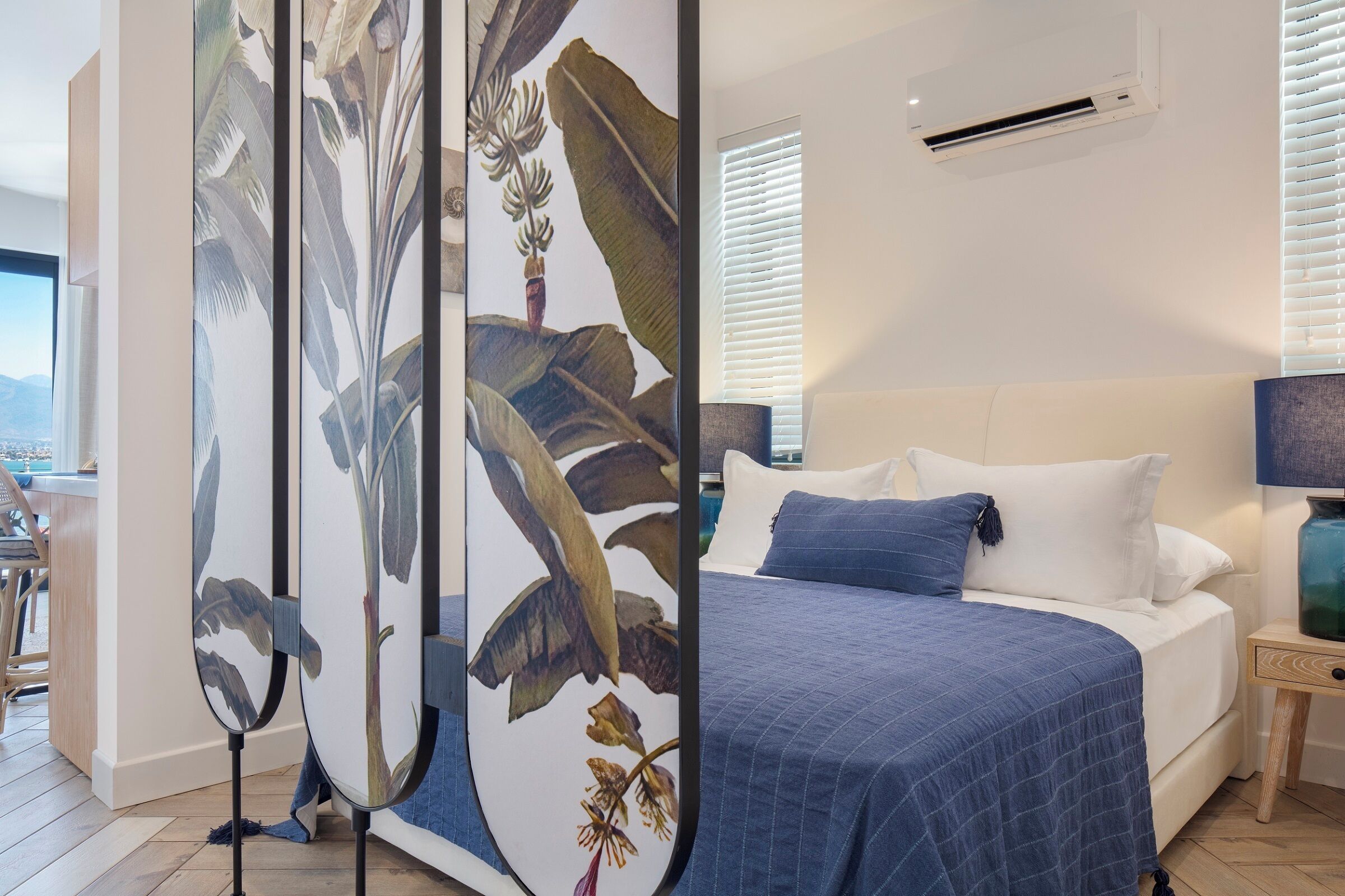 A bedroom with a bed, white pillows, a blue bedspread, a nightstand with a lamp, and a wall-mounted air conditioner.