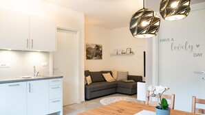 Una cocina moderna con armarios blancos, una mesa de comedor de madera y una sala de estar con sofá y decoración mural.