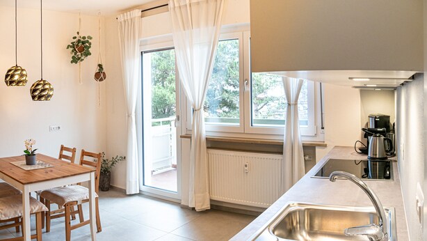 Una cocina moderna con un lavamanos, estufa y cafetera. Hay una mesa de comedor con sillas y un ventanal con cortinas.