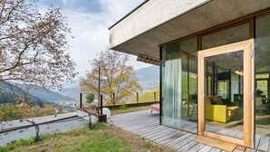 Una casa moderna con una terrazza affacciata su un fiume, una porta in vetro che dà su un divano giallo e vista sulle montagne.