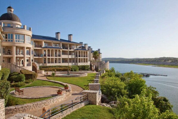 A large, multi-story hotel with a central courtyard, a fountain, and a view of a lake. - Sweeping Views of Lake Travis, Luxury Resort Amenities with 3 Pools, Hot Tub and Fitness Center (Lago Vista)