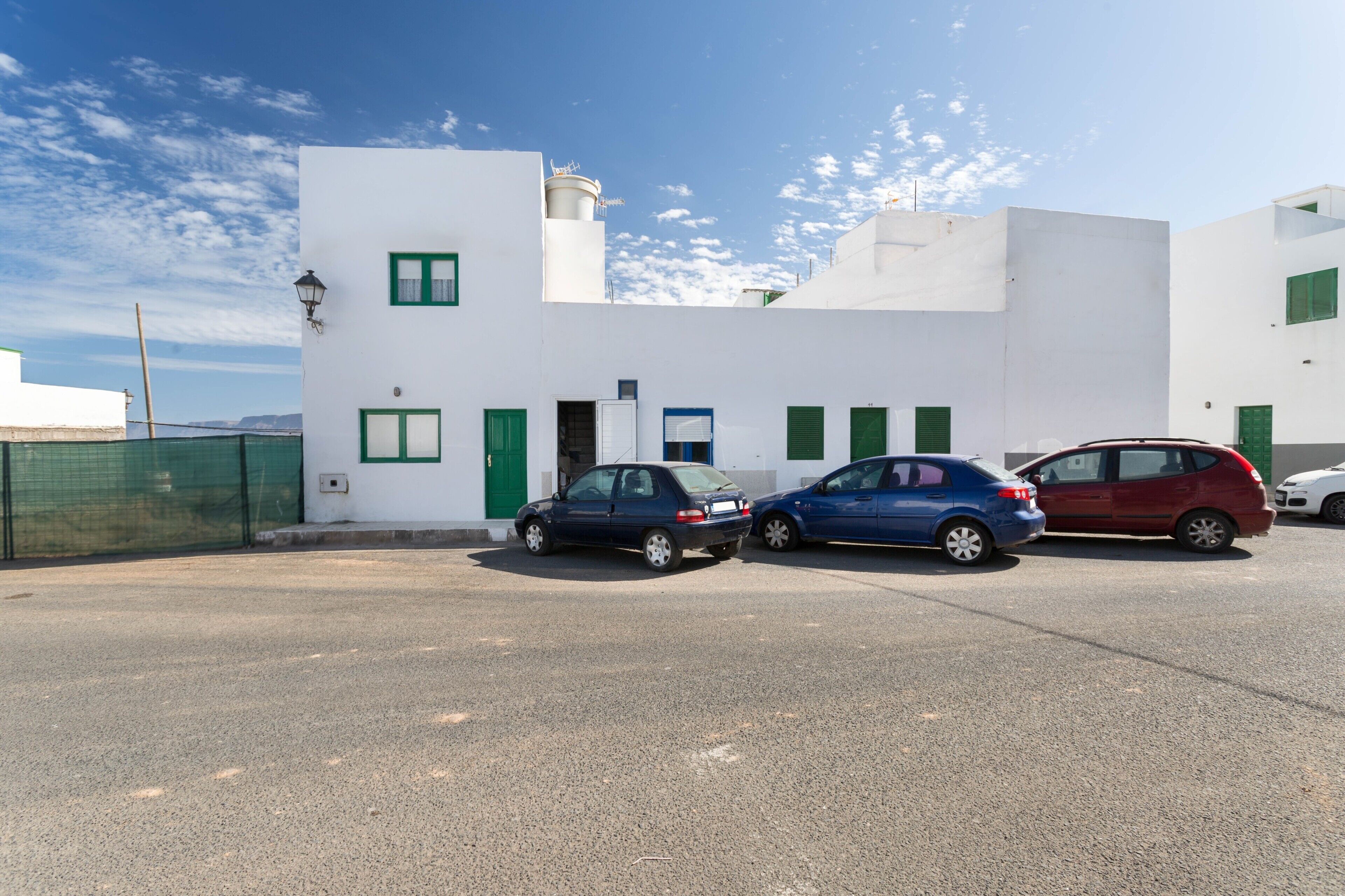 A white building with green shutters, a blue car, and a red car parked in front.