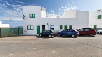 A white building with green shutters, a blue car, and a red car parked in front.