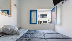 Un dormitorio con una cama, una ventana de marcos azules, un televisor montado en la pared y un espejo.