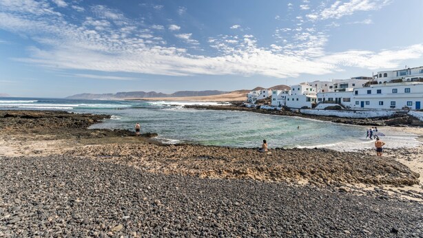 Zona costera con edificios blancos, playa rocosa y cielo azul despejado.