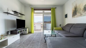 Una sala de estar moderna con televisor de pantalla plana, sofá gris, mesa de centro de vidrio y un balcón con vistas.