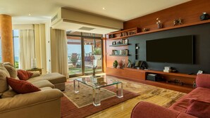A modern living room with a glass coffee table, beige sofas, a flat-screen TV, wooden shelves with decorative items, and a large window offering a view. - Villa "Albella" with Sea View, Garden, Terraces, Pool, Sauna, Whirlpool & Gym (San Bartolone de Tirajana)