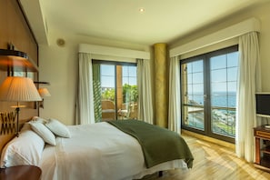 A hotel room with a bed, a chair, a table, a lamp, and large windows with a view of the sea.