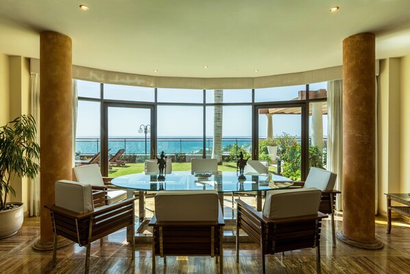 A spacious room with a large glass table surrounded by chairs, large windows offering a view of the sea, and a potted plant.