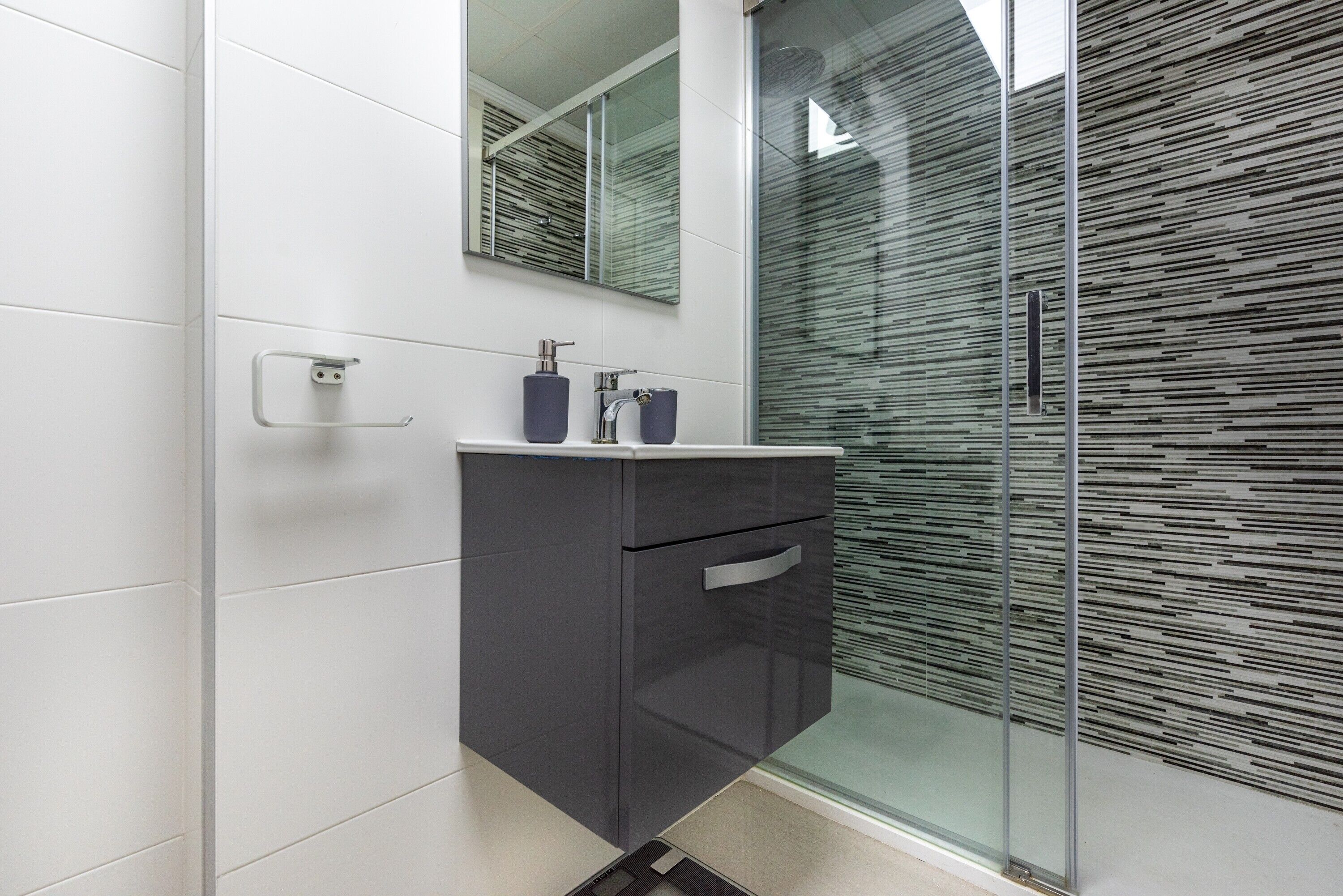 Un baño moderno con cabina de ducha de vidrio, tocador blanco con mueble oscuro y espejo sobre el lavabo.