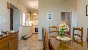 Un comedor con mesa de madera, sillas, un jarrón de flores y una cocina al fondo.