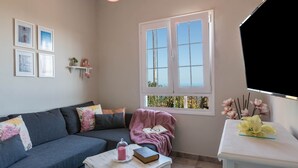 Un salón con sofá gris, una mesa de centro blanca con libros y un vaso, una ventana con vistas, y un estante blanco con adornos.