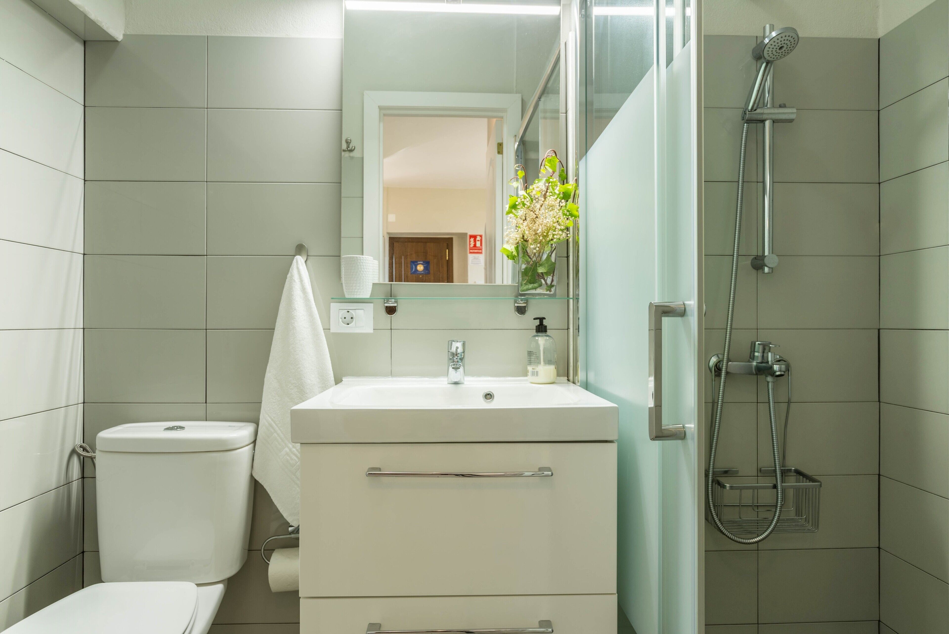 Un baño con inodoro, lavamanos, espejo, ducha y un pequeño florero con flores.