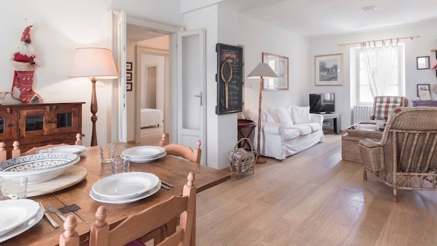 Un comedor con una mesa de madera dispuesta para cuatro personas, un sofá blanco, sillas de mimbre y un televisor al fondo.