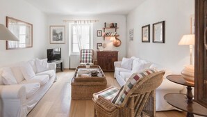 Una sala de estar con dos sofás blancos, un sillón de mimbre, una mesa de centro, un televisor y una estantería.