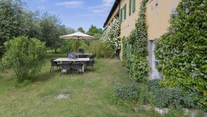 Eine Terrasse mit Tisch und Stühlen, umgeben von viel Grün und einem Gebäude mit grünen Fensterläden.
