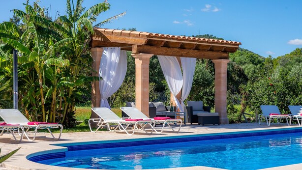 Ein Poolbereich mit Liegestühlen, einer Pergola mit weißen Vorhängen und üppiger Bepflanzung.