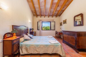 A bedroom with a bed, bedside table, lamp, dresser, window, and a view of greenery outside.