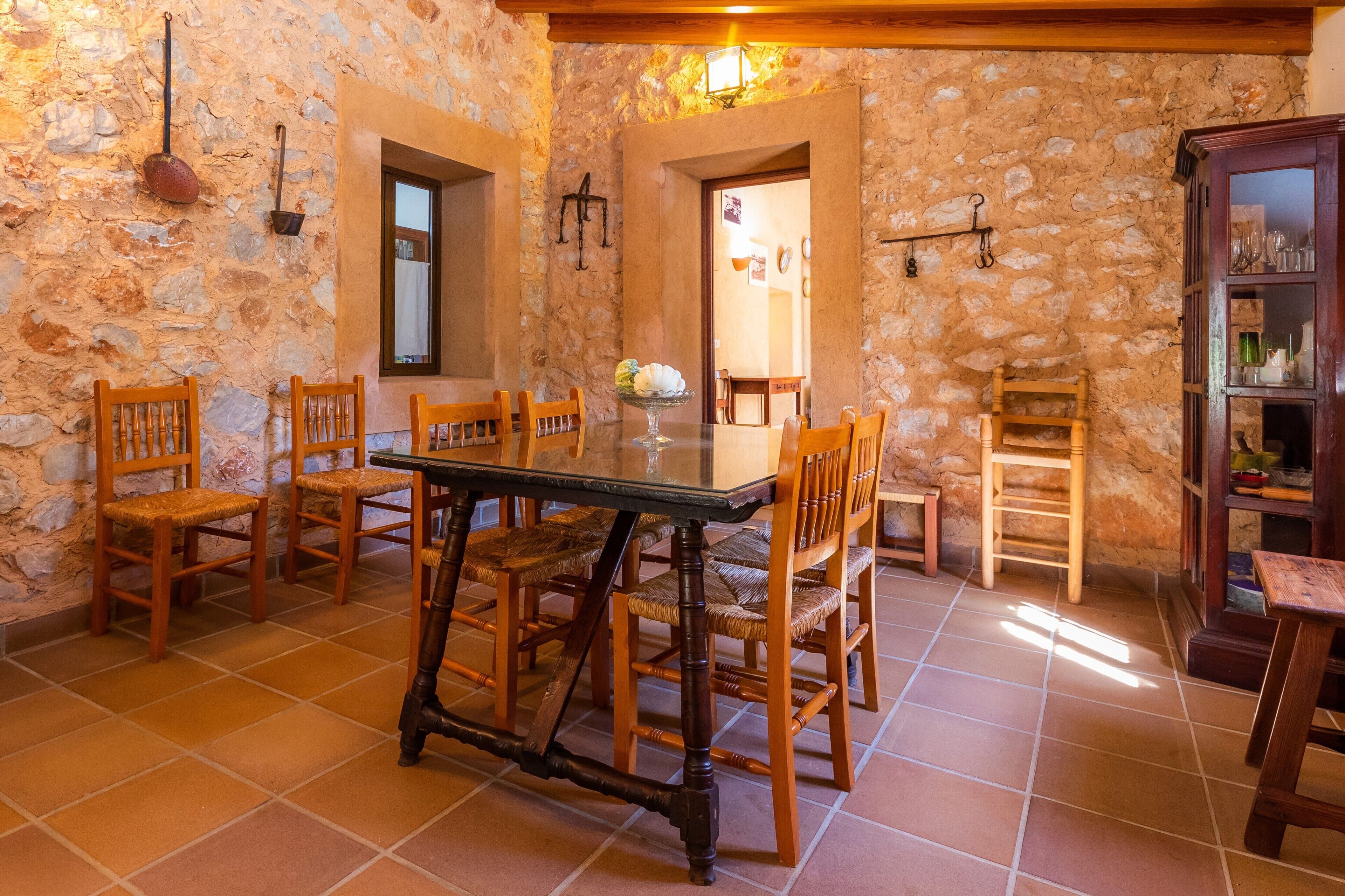 A stone-walled dining area with a wooden table and chairs, a tiled floor, and a wooden cabinet with glass doors.