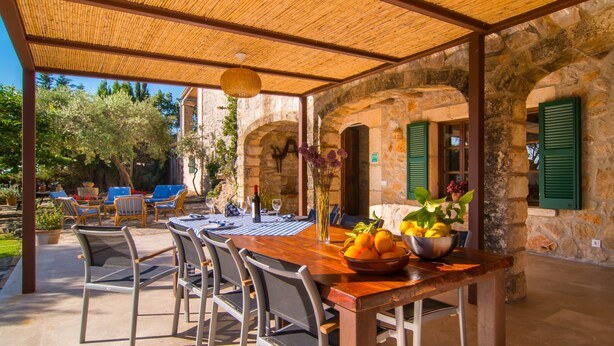 An outdoor dining area with a stone wall, a wooden table, metal chairs, and a basket of fruit. - Stunning Holiday Home “Finca Rafal” with Garden, Pool, Terraces & WiFi; Parking Available (Son Servera)