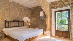 A bedroom with a stone wall, a wooden bed, a nightstand, a lamp, and a view through a door to an outdoor area. - Stunning Holiday Home “Finca Rafal” with Garden, Pool, Terraces & WiFi; Parking Available (Son Servera)