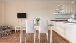 A dining area with a glass table and white chairs, a flat-screen TV mounted on the wall, and a kitchen area with white cabinets and a tiled backsplash.