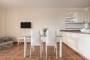 A dining area with a glass table and white chairs, a flat-screen TV mounted on the wall, and a kitchen area with white cabinets and a tiled backsplash. - Holiday Apartment “Carla 1A” in Playa San Juan with Sea and Mountain Views (Playa San Juan)
