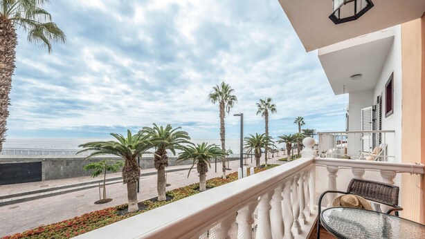 A balcony with a view of the sea, palm trees, and a tiled floor.