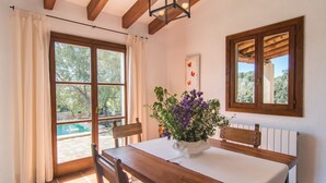 Un coin repas avec une table en bois, des chaises, un vase de fleurs, une fenêtre donnant sur un paysage verdoyant et un plafond avec des poutres apparentes.