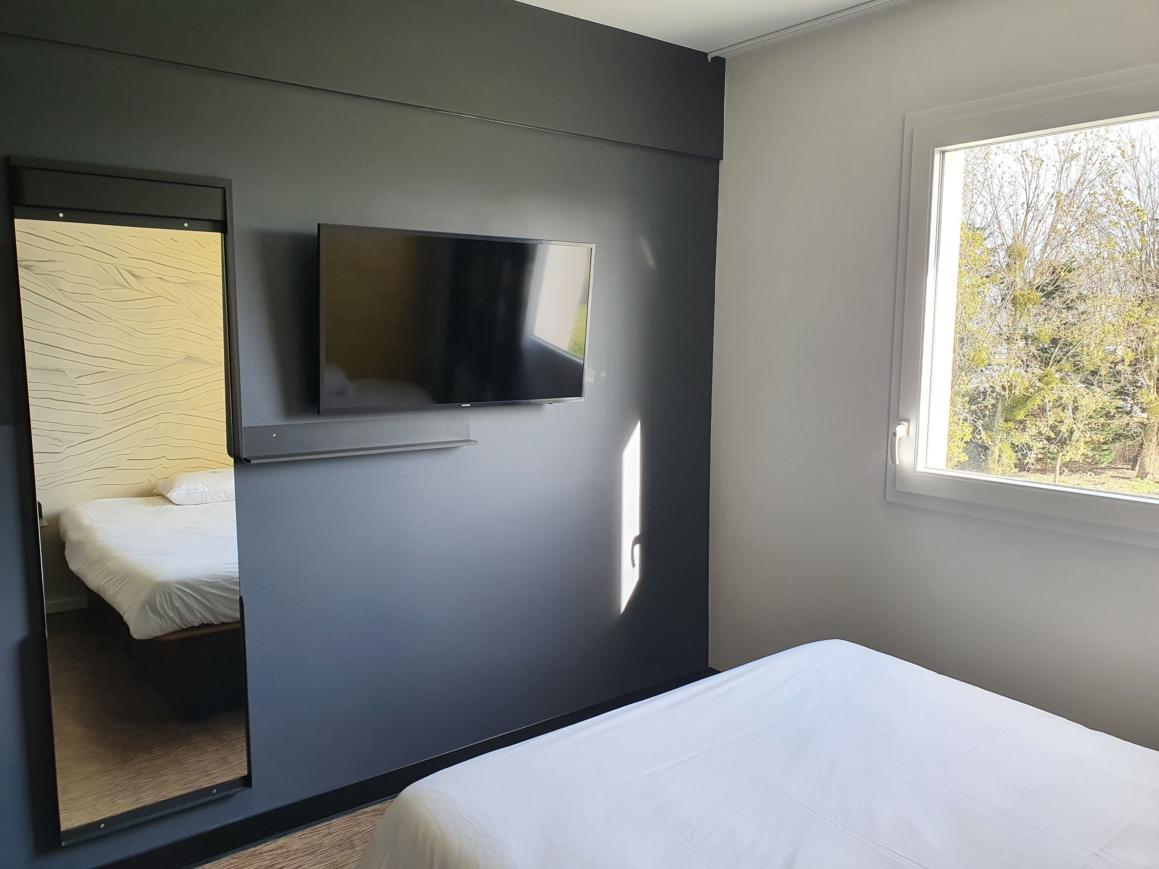 A bedroom with a bed, a wall-mounted TV, and a window with a view of trees.