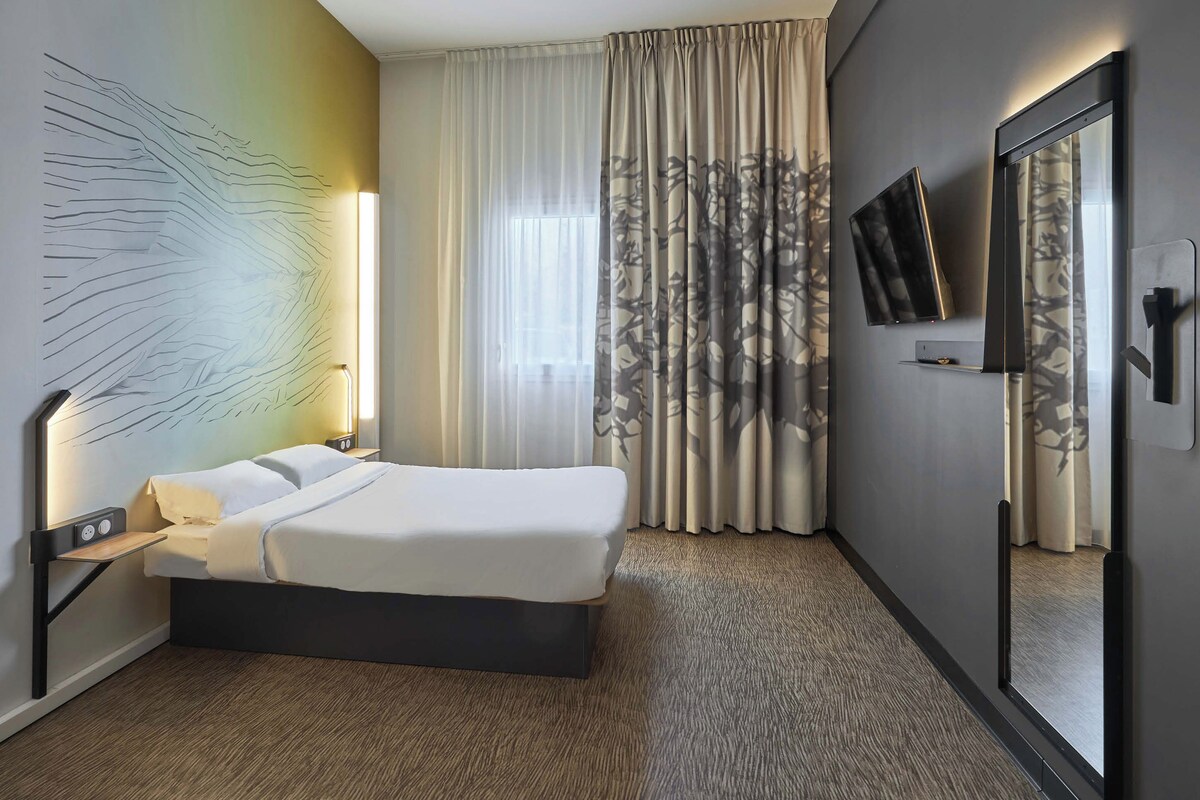 Una habitación de hotel moderna con una cama grande, una mesita de noche con lámpara, un televisor montado en la pared y un espejo amplio.