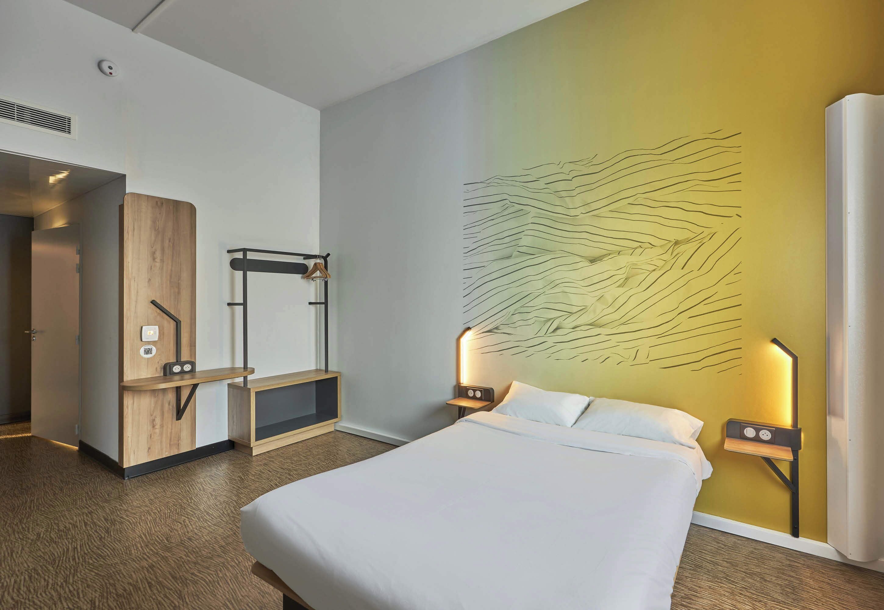 Habitación de hotel con cama, escritorio y lámpara fijada a la pared.