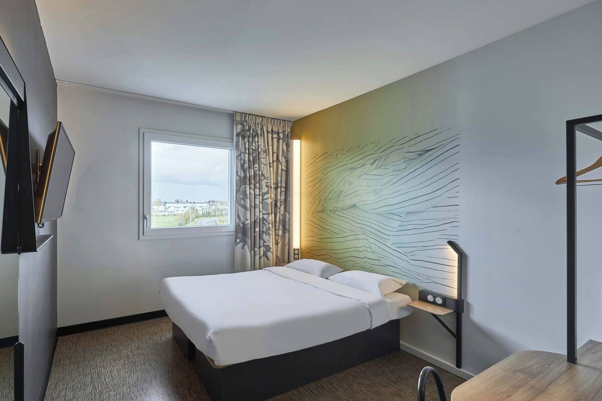 A hotel room with a bed, a desk, a chair, a window, and a wall-mounted light fixture.