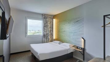 A hotel room with a bed, a desk, a chair, a window with curtains, and a wall-mounted light fixture.