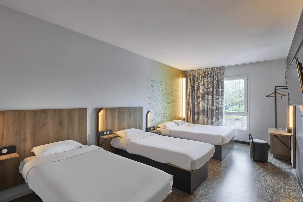 A hotel room with three single beds, a wooden headboard, a bedside table with a lamp, a window with curtains, and a small stool.