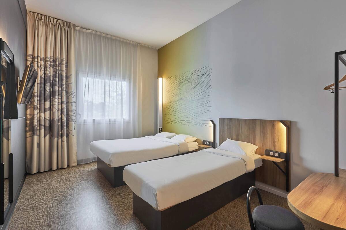 A hotel room with two beds, a desk, a chair, a lamp, and a large window with curtains.