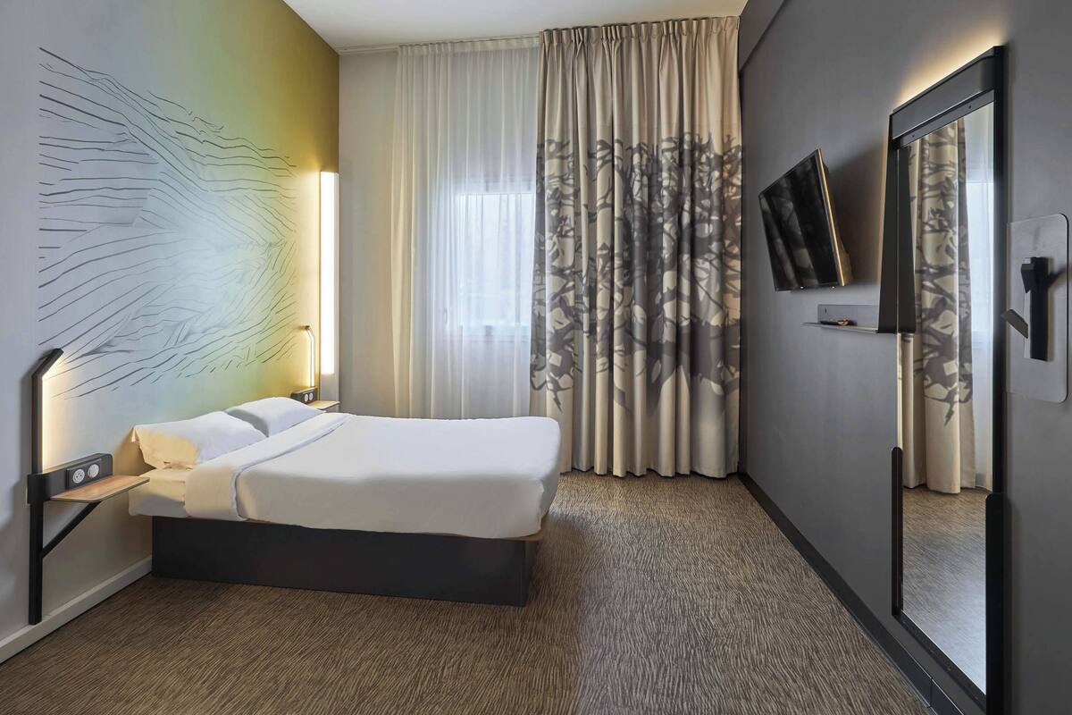 A modern hotel room with a large bed, a bedside table with a lamp, a wall-mounted TV, and a large window with sheer curtains.
