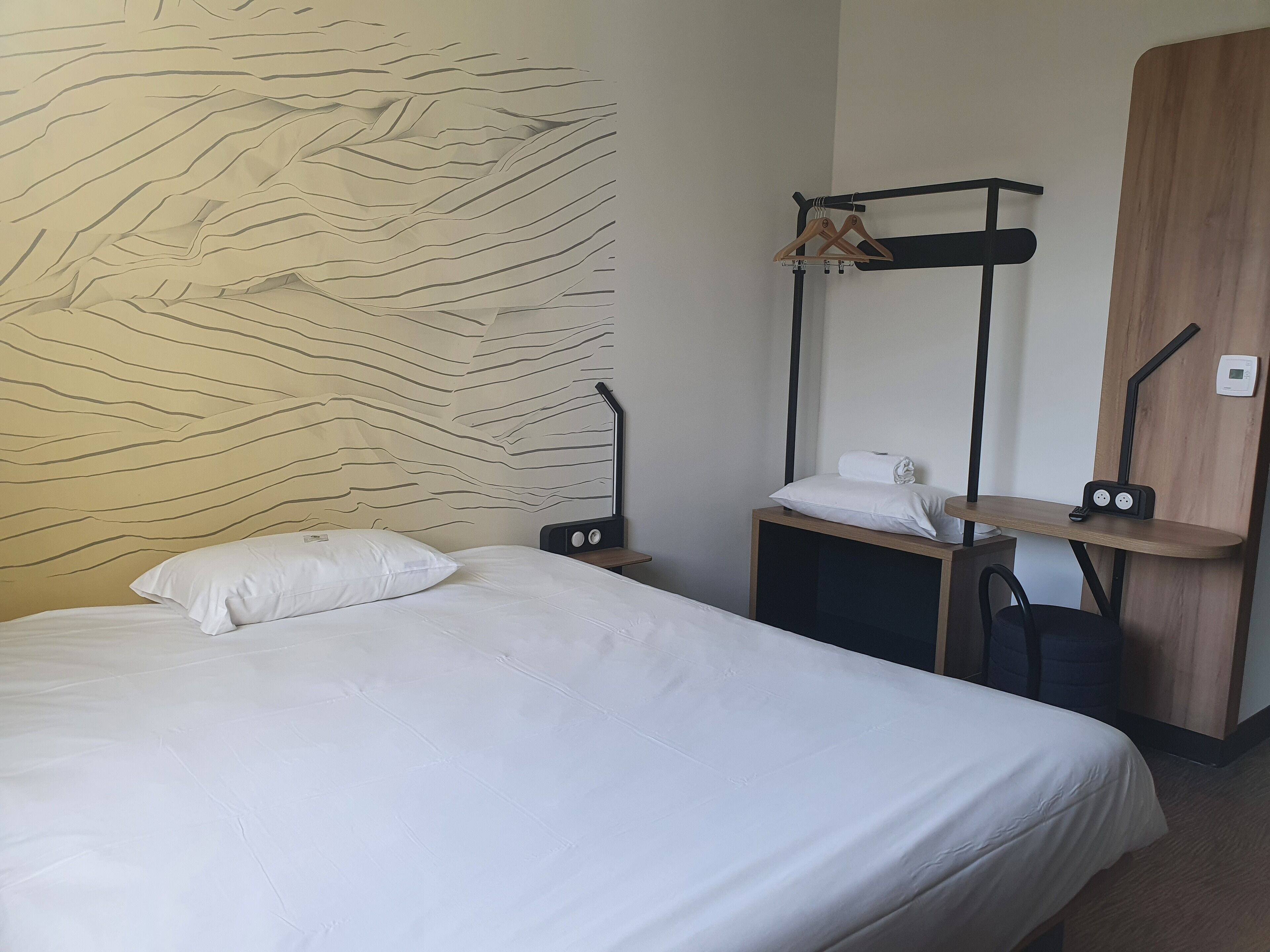 Quarto de hotel com cama, mesa de cabeceira de madeira, prateleira na parede com uma toalha e uma pequena mesa, e uma parede com textura.
