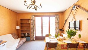 A cozy room with a sofa, a dining table set for four, a bookshelf, and a television mounted on the wall.