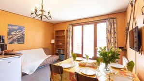 A compact room with a bed, a small desk, a dining table set for two, and a large window with curtains.