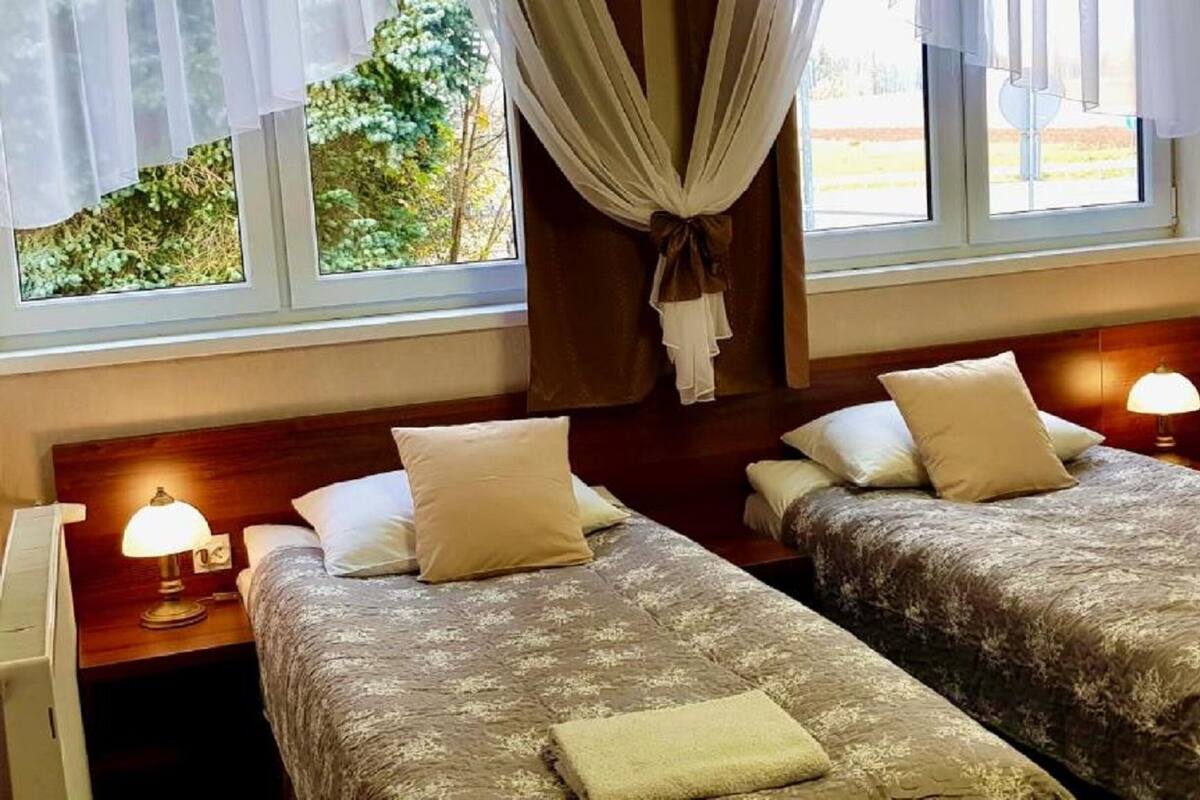 A hotel room with two beds, each with a pillow and a blanket. There are bedside lamps and a window with curtains.