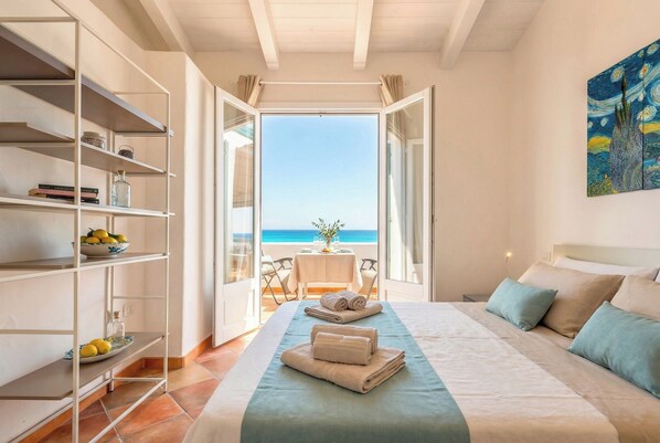 A hotel room with a bed, a bookshelf, a table with chairs, and a view of the sea.