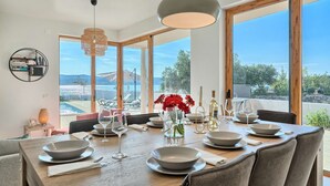 Comedor con mesa de madera dispuesta para seis personas, ventanas amplias con vistas al mar y una lámpara colgante moderna.