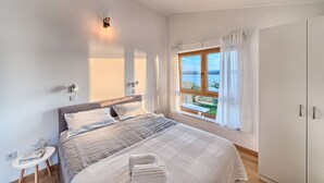 Un dormitorio con una cama grande, una mesita de noche con lámpara, una ventana con cortinas y vistas al mar.