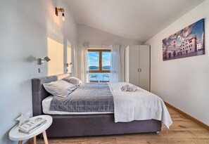 A bedroom with a large bed, a bedside table with a book, a window with curtains, and a painting on the wall.