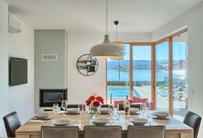 A dining area with a table set for six, a television on the wall, and a view of a lake through large glass doors.