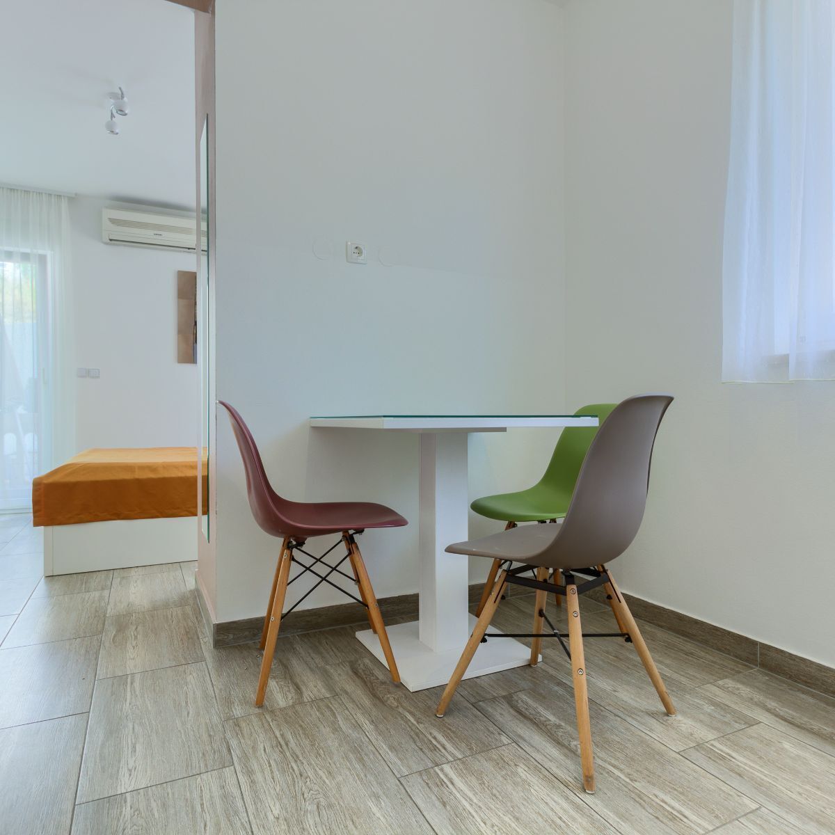 Une pièce moderne avec une table à plateau en verre, deux chaises aux pieds en bois, un banc et un climatiseur blanc fixé au mur.